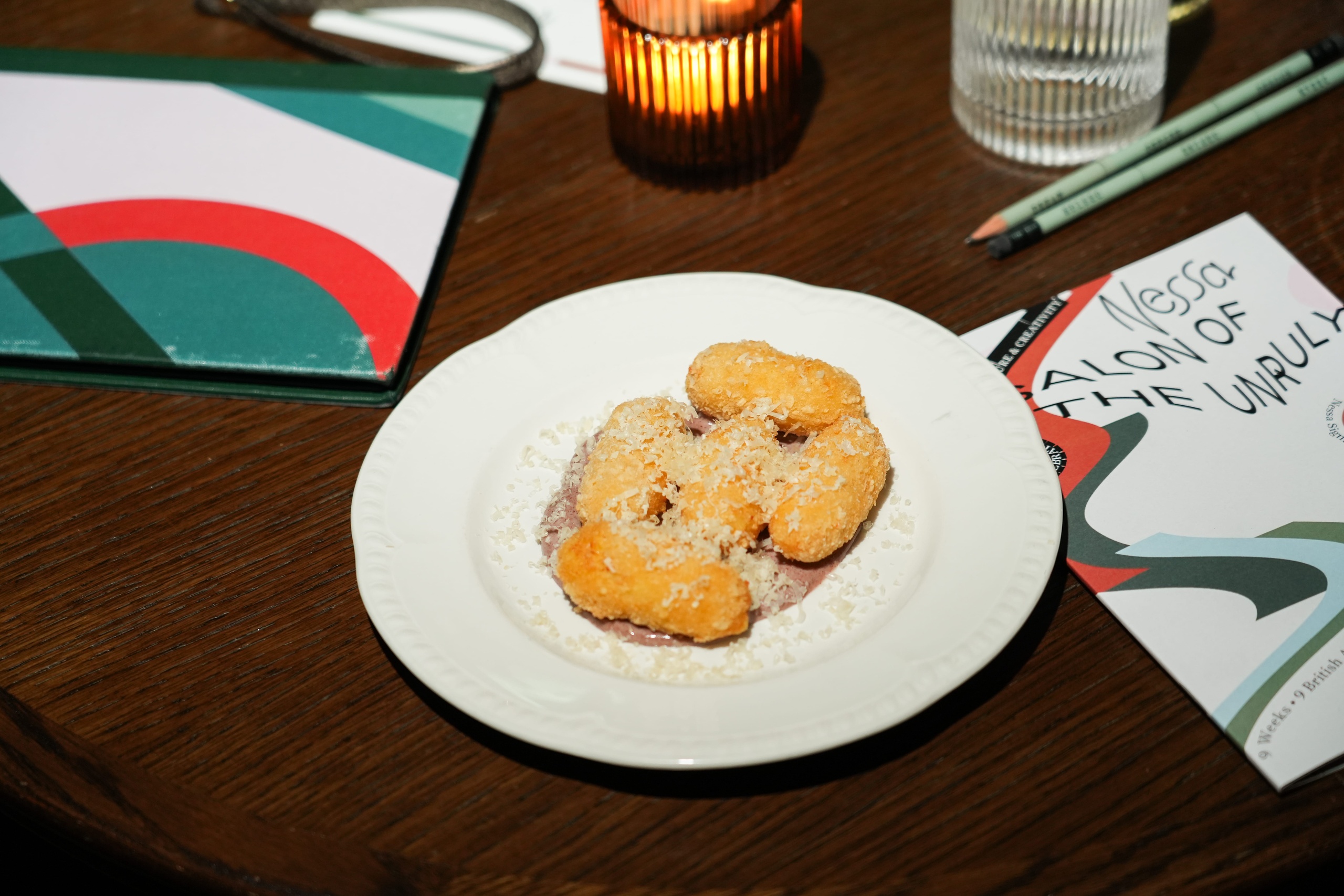 Plate of crispy croquettes on creamy sauce next to Nessa’s branded “Nessa Salon of the Unruly” menu and lit candle on a dark wood table