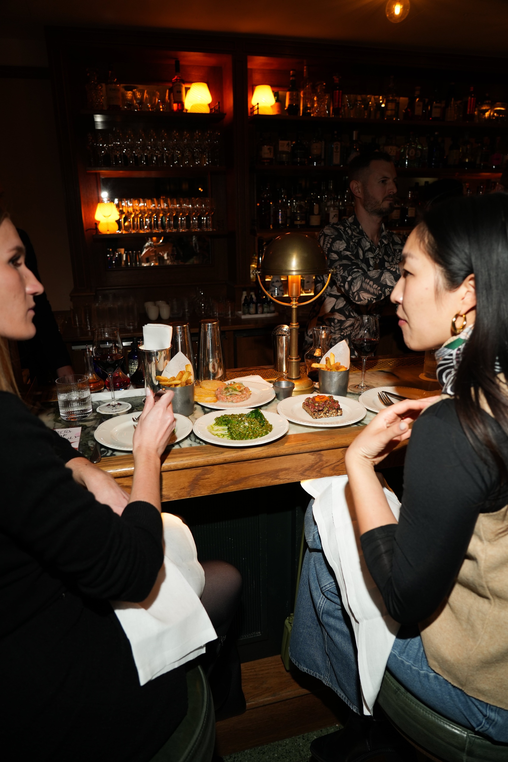 Two women dining at the bar in Nessa Soho, enjoying tartare, greens, and fries under warm lighting with a bartender in the background