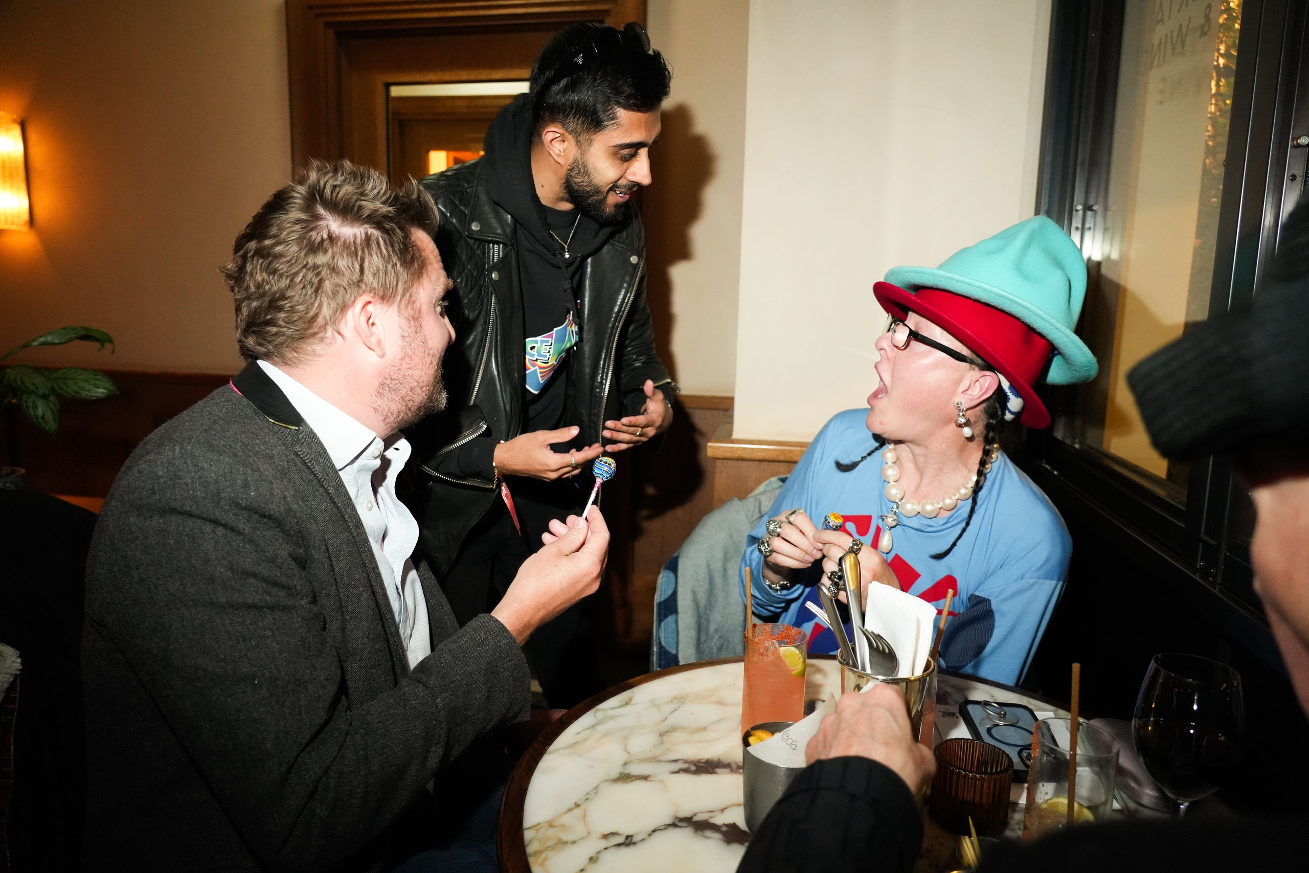 Three people laughing and talking animatedly at a round marble table in Nessa, Soho restaurant, with one guest wearing bold fashion and layered hats