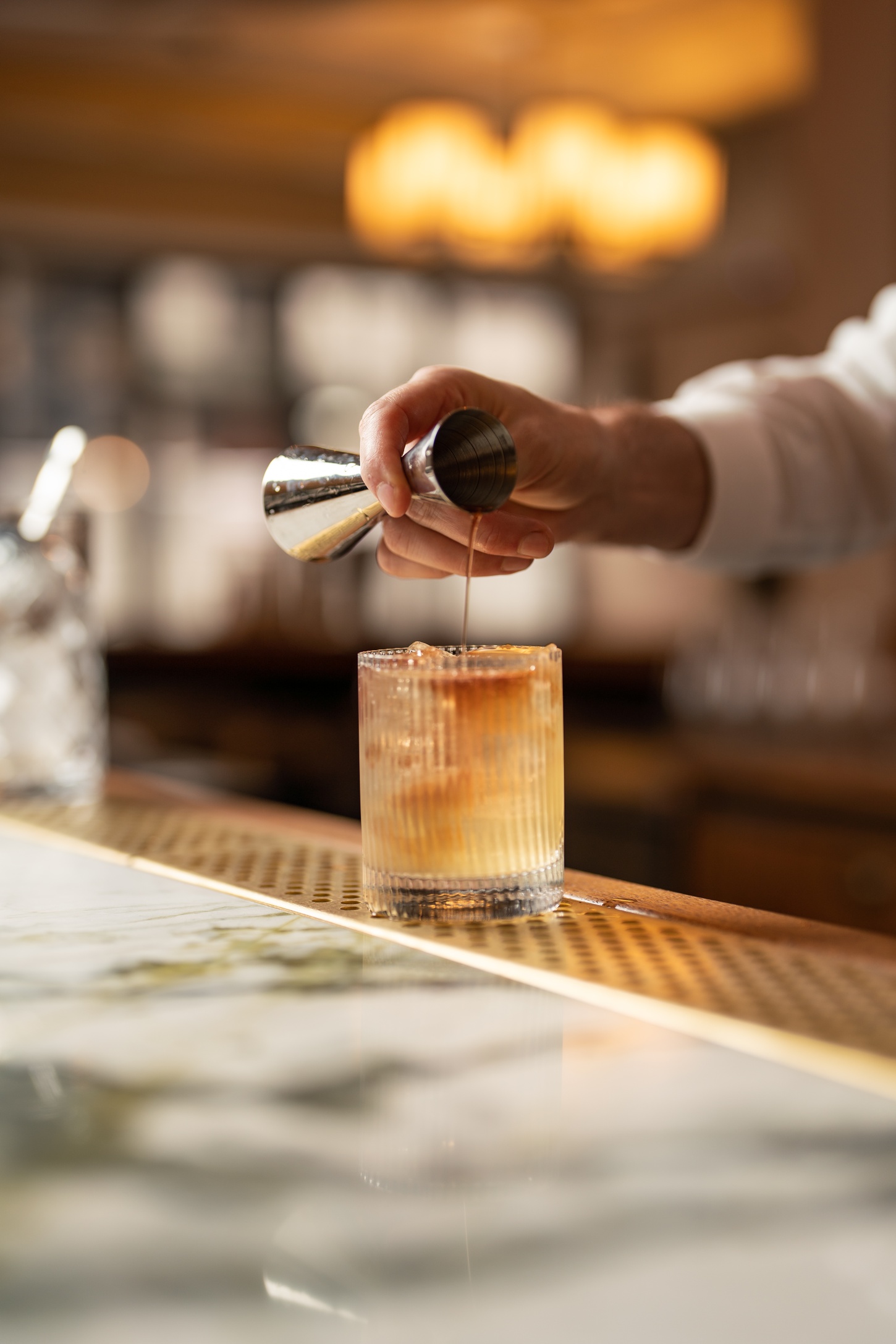 Close-up of a bartender pouring a cocktail from a jigger into a rocks glass on the marble bar at Nessa restaurant in Soho