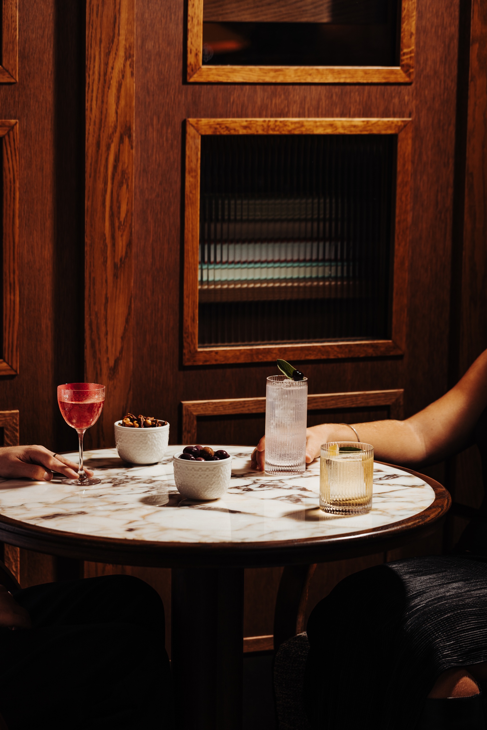 Two guests enjoying cocktails, olives, and spiced nuts at a round marble table with wooden panel backdrop at Nessa restaurant in Soho
