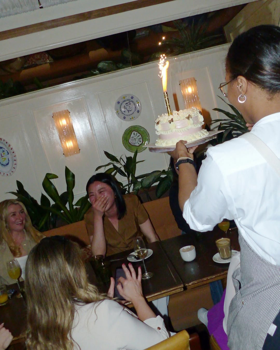 Server delivering a birthday cake with sparkler candles to a laughing group of women at a warmly lit table at Nessa restaurant in Soho