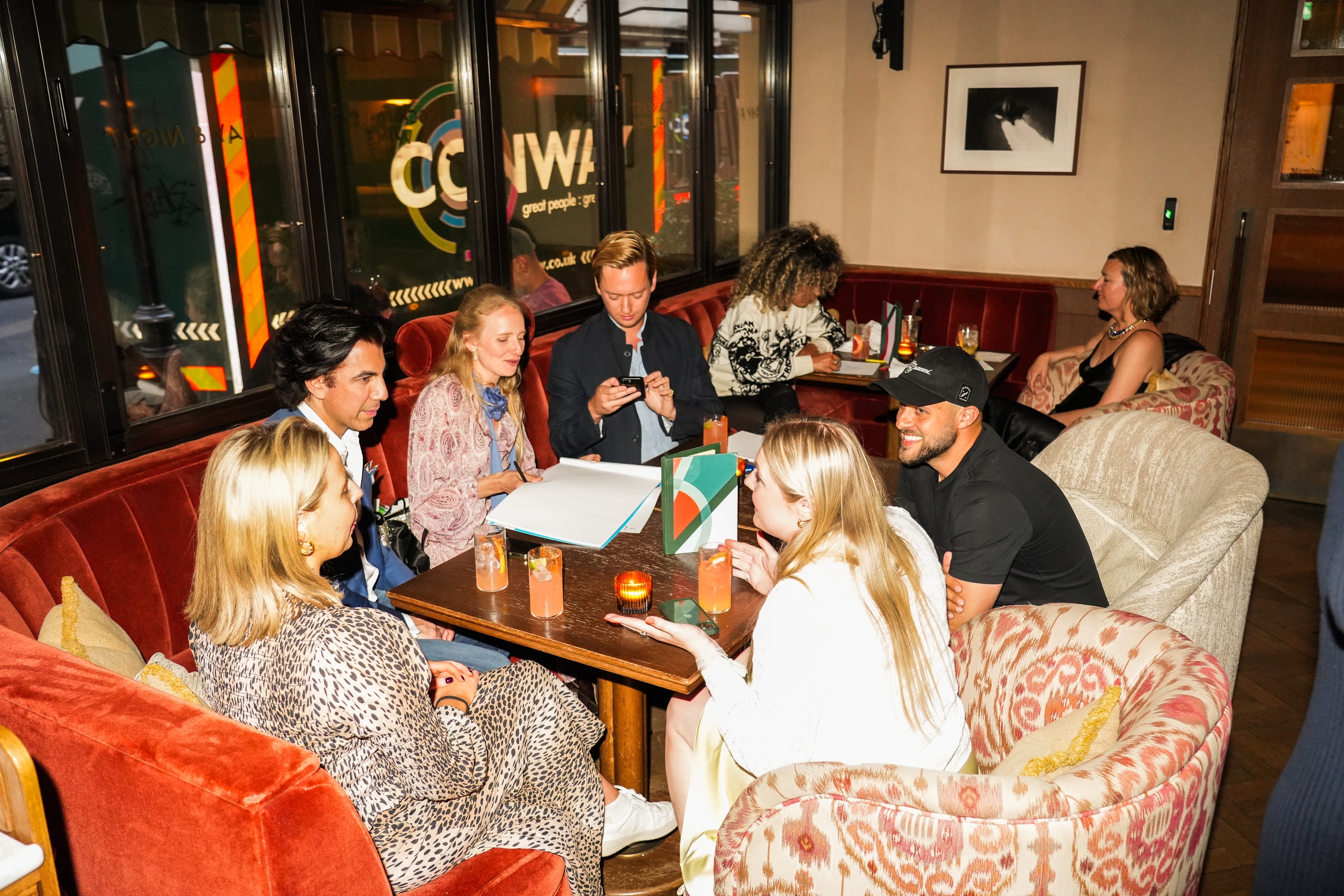 A lively group seated in a cozy lounge area at Nessa, enjoying cocktails, conversation, and collaborative sketching in a warmly lit, velvet-lined corner of the restaurant.