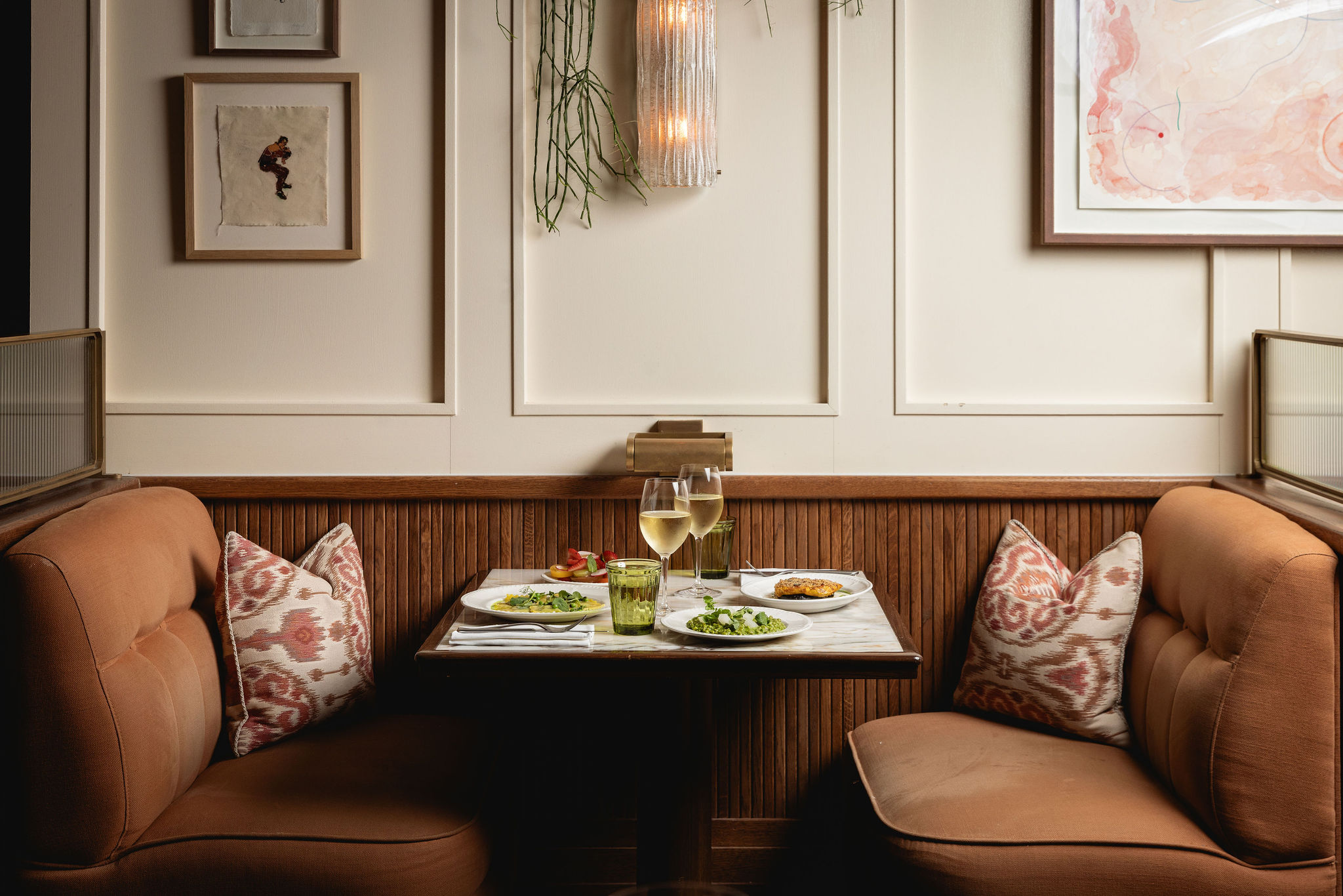 Cozy booth for two with a marble table, plates of food, and white wine surrounded by art and plants at Nessa restaurant in Soho