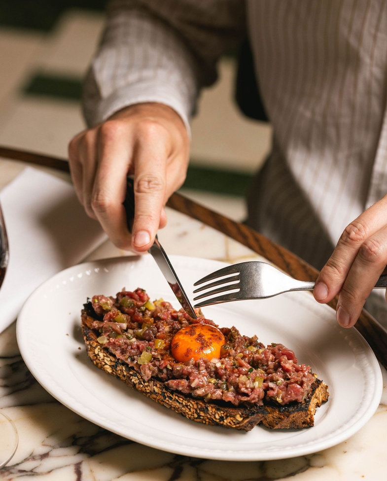 Close-up of a diner slicing into beef tartare on seeded sourdough at Nessa, Soho — topped with a golden egg yolk, served on a marble table.