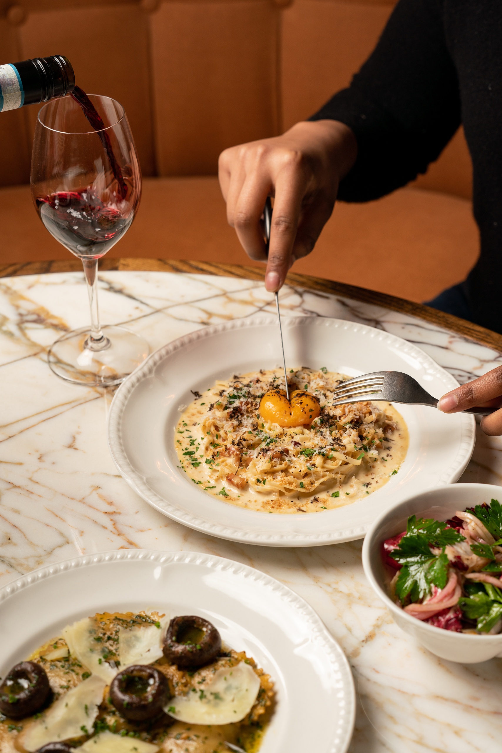 Close-up of a diner slicing into egg-topped creamy tagliatelle with a glass of red wine being poured at Nessa restaurant in Soho