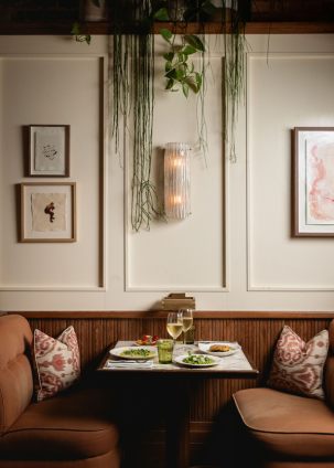 A cosy table for two at Nessa in Soho, set with small plates and wine, framed by blush banquettes, soft lighting, trailing greenery, and modern art.