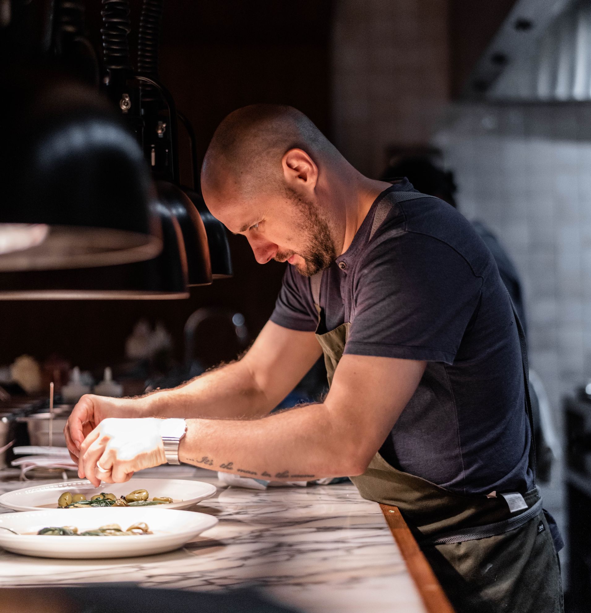 om Cenci, Executive Chef at Nessa, plating a dish under kitchen lights in Soho restaurant.