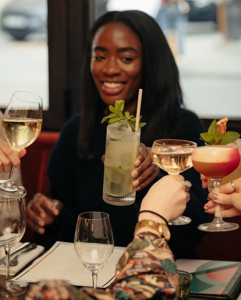 Group of friends toasting with cocktails and wine at a restaurant table, smiling woman in focus