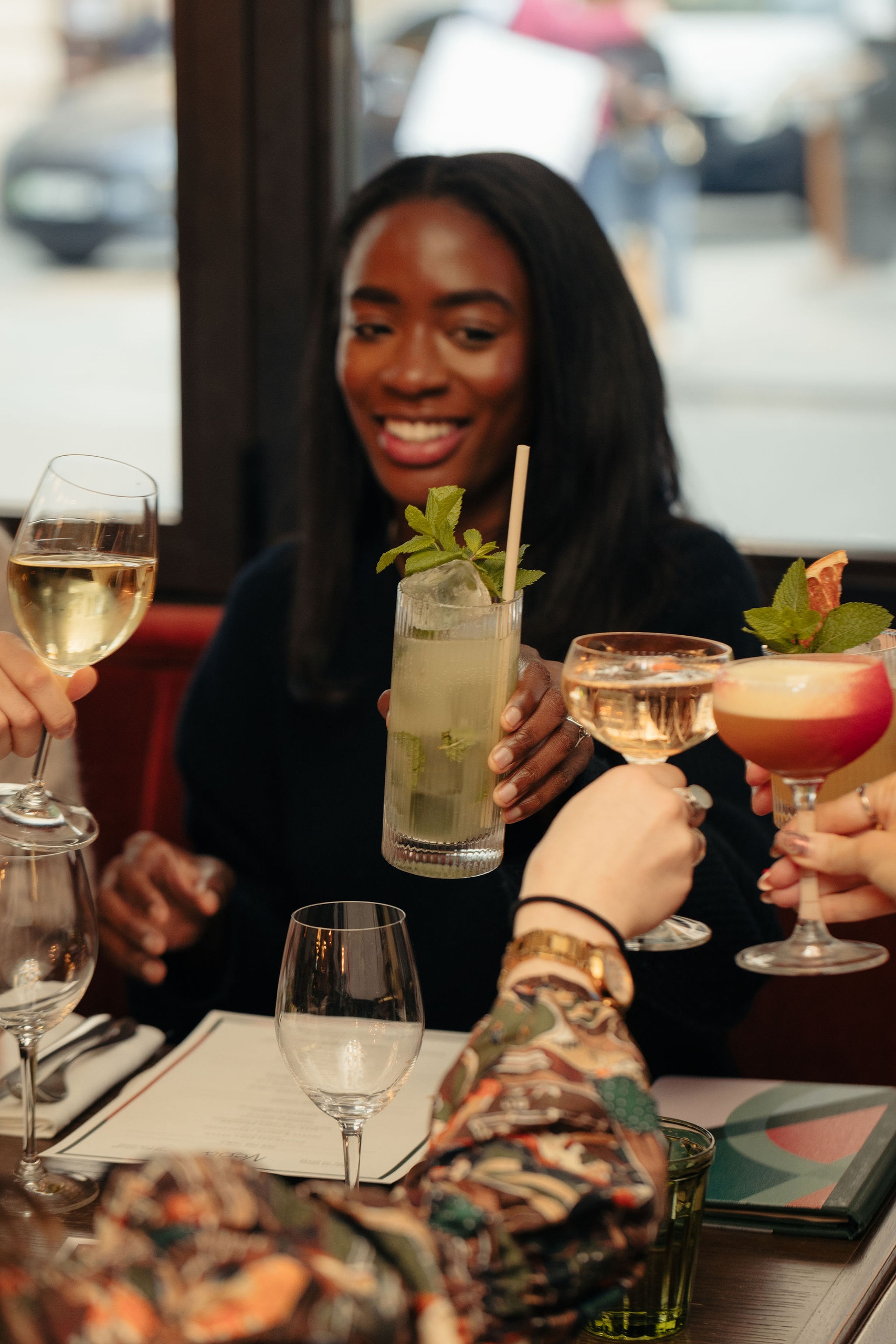 Group of friends toasting with cocktails and wine at a restaurant table, smiling woman in focus