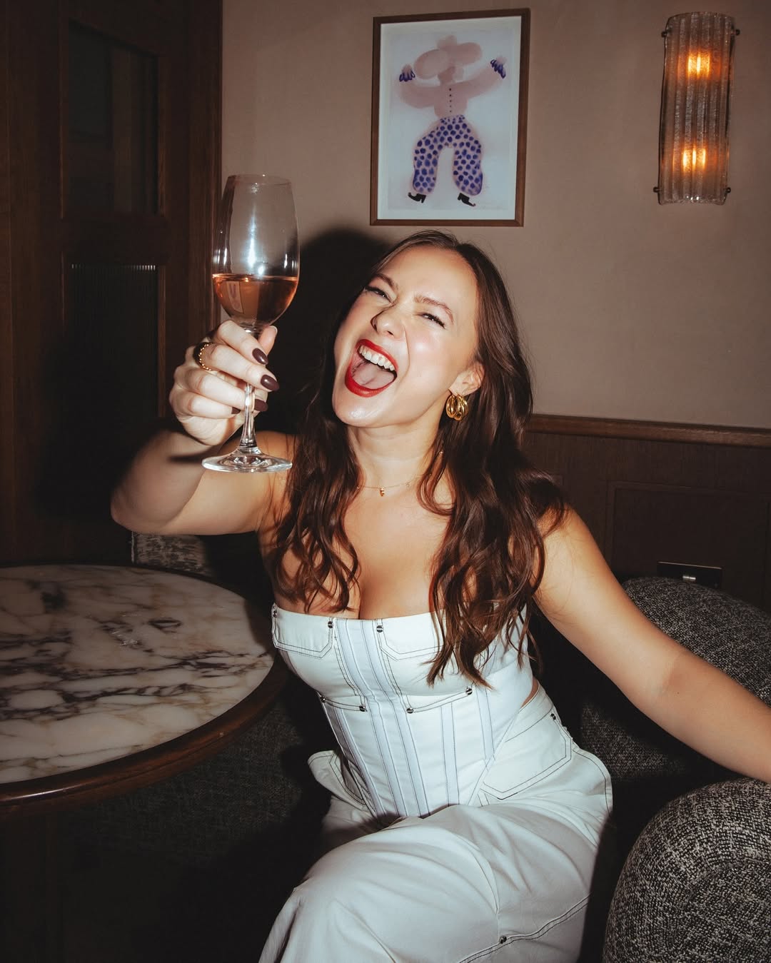 Smiling woman in a white corset top holding up a glass of rosé wine in a stylish lounge